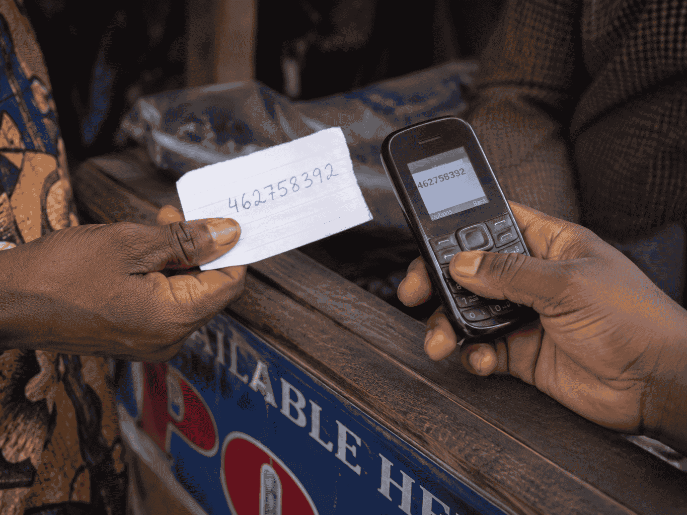 A handwritten 9-digit Ticket IT code on a paper slip, being keyed into a basic feature phone at a stall